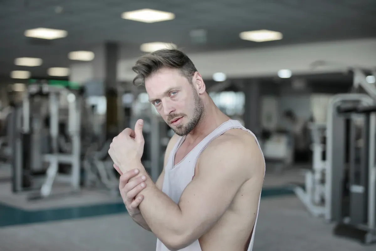 Side view of strong male athlete in casual sportswear standing with raised crossed hands near chest while resting after workout in modern fitness center and loo