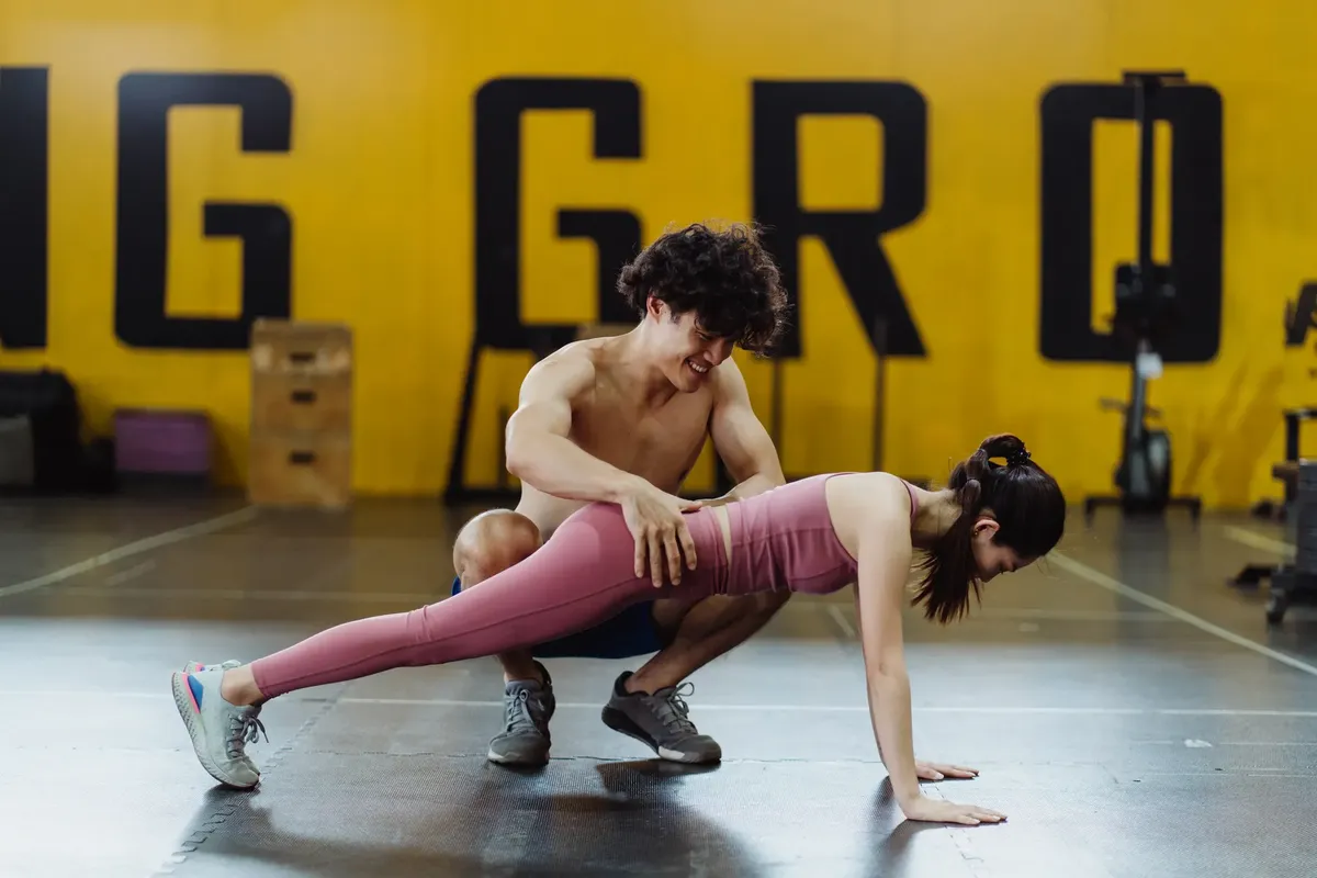Personal trainer helps woman with push-up exercise in gym setting, promoting fitness.