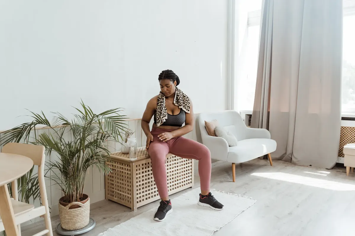 Black woman in activewear resting at home after exercise, sitting on a chair with a towel around her neck.
