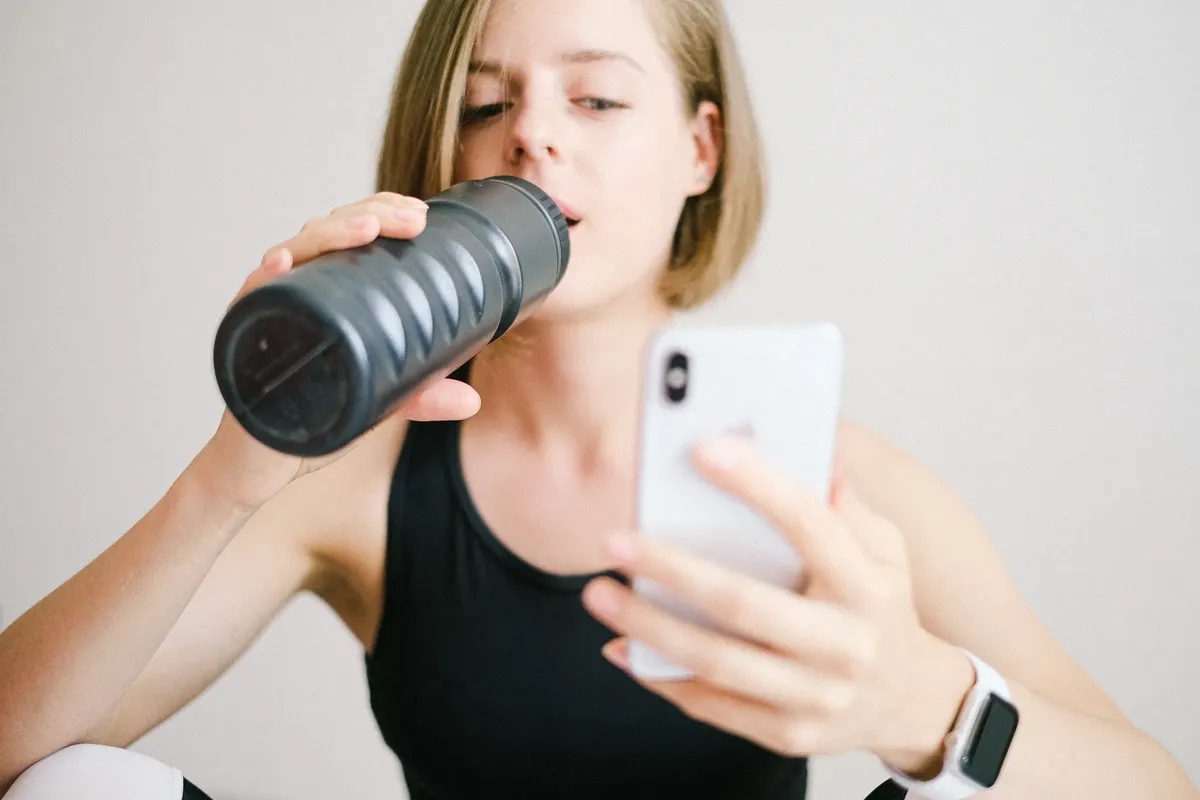 Woman drinking water while using a smartphone in a home fitness setting, highlighting modern technology and hydration.